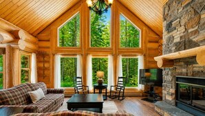 A log cabin interior with a fireplace, large windows, a plaid sofa, and a dining table. - Fiddler Lake Resort - Rabbit 24 (Mille-Isles)