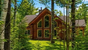 A log cabin with a large central window, surrounded by dense forest. - Fiddler Lake Resort - Rabbit 24 (Mille-Isles)