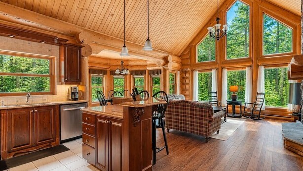 A spacious log cabin interior with a kitchen, dining area, and large windows offering a view of the forest. - Fiddler Lake Resort - Rabbit 24 (Mille-Isles)