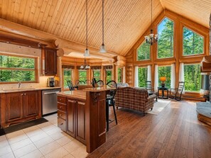 Ein geräumiges Blockhaus-Interieur mit Küche, Essbereich und großen Fenstern, die einen Blick auf den Wald bieten.