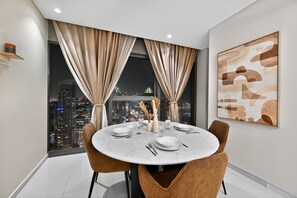 A modern dining room with a round table set for four, large windows with curtains, a cityscape view, and a piece of abstract art on the wall. - Burj & Sea Views |Luxury 2BR| 5-Star Amenities + Concierge services (Dubai)