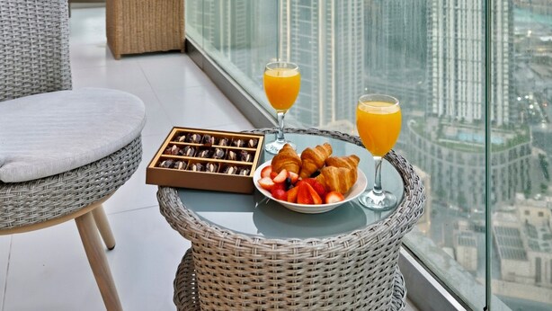 Ein Balkon mit Korbmöbeln, einem Frühstücksbrett mit Croissants, Erdbeeren und Orangensaft, sowie Blick auf den Burj Khalifa.