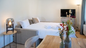 Une chambre d’hôtel moderne avec un lit bien fait, une petite table ornée d’un vase de fleurs, une chaise, une table de chevet avec une lampe et un téléviseur fixé au mur.