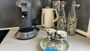 Keittiön työtasolla on kahvinkeitin, vedenkeitin, kaksi lohikäärmekuvioitua mukea, lautanen, jossa on kaksi teepussia, ja koristeellinen pullo.