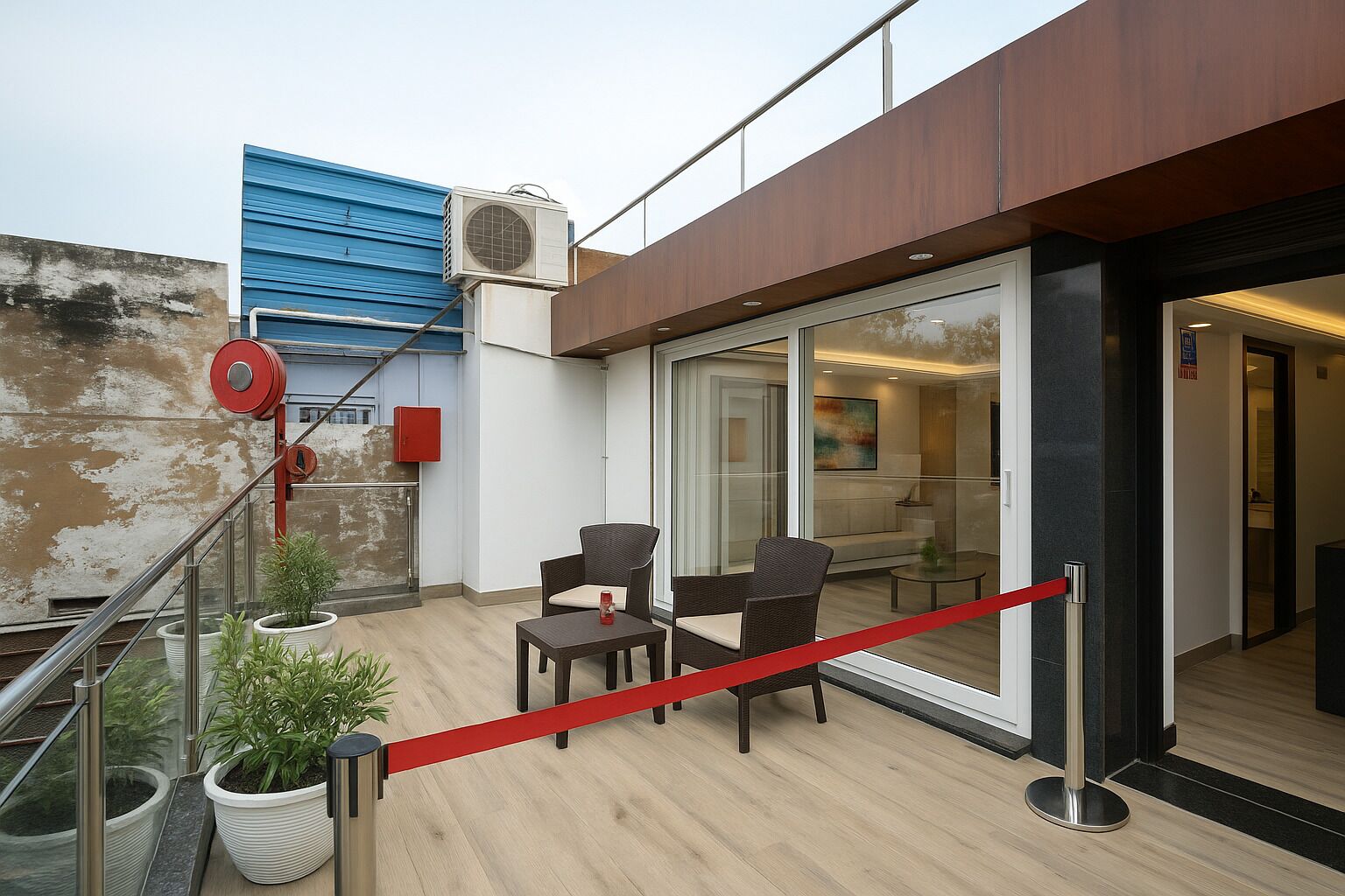 Un espacio exterior moderno con dos sillas y una mesa, una barrera de cuerda roja y un edificio azul al fondo.