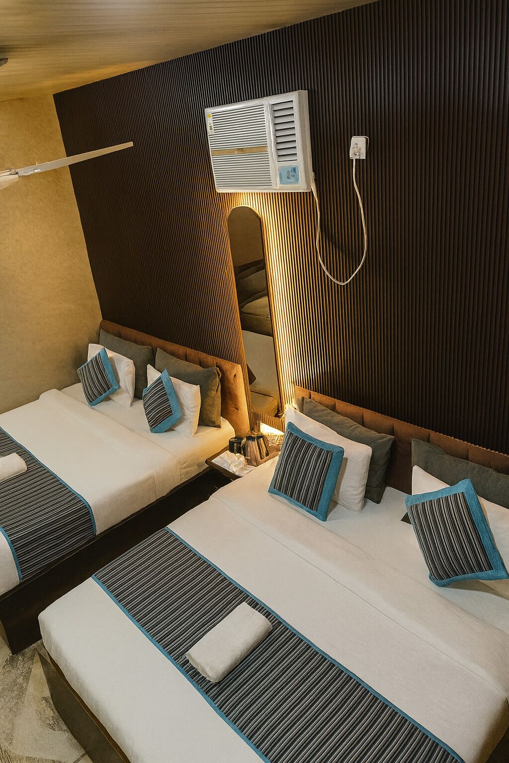 A hotel room with two single beds, a wooden headboard, a wall-mounted air conditioner, and a bedside table with a lamp.