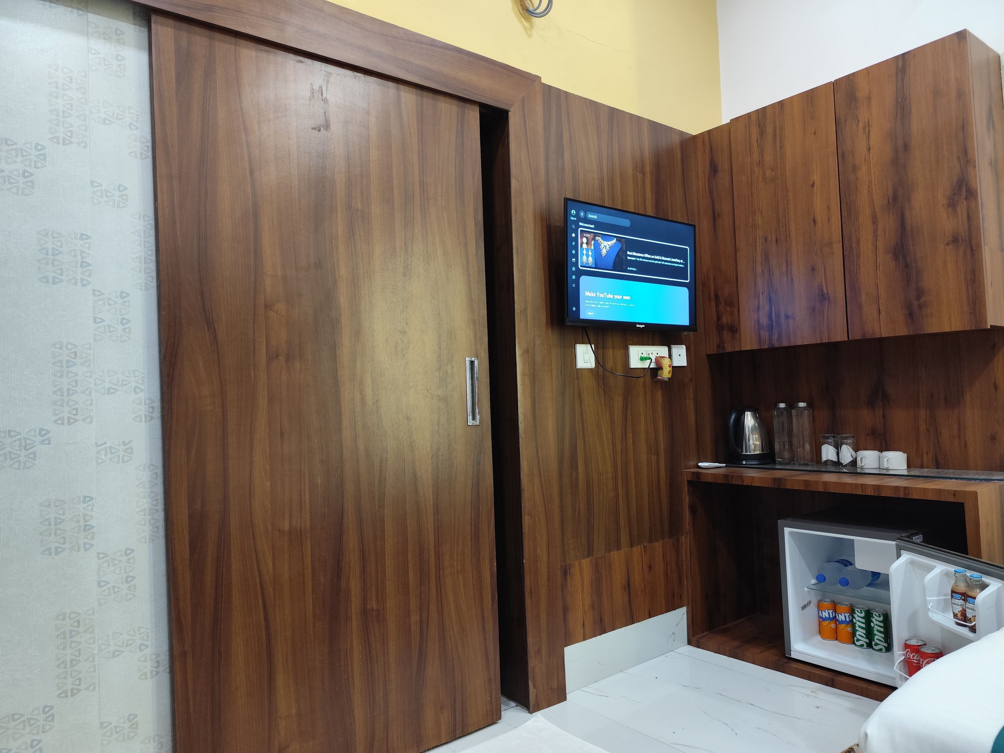 Una habitación con armarios de madera, un televisor montado en la pared y un refrigerador lleno de bebidas.