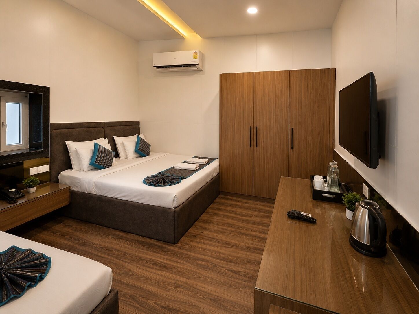 A modern hotel room with a bed, bedside table, TV, wardrobe, and a desk with a kettle.