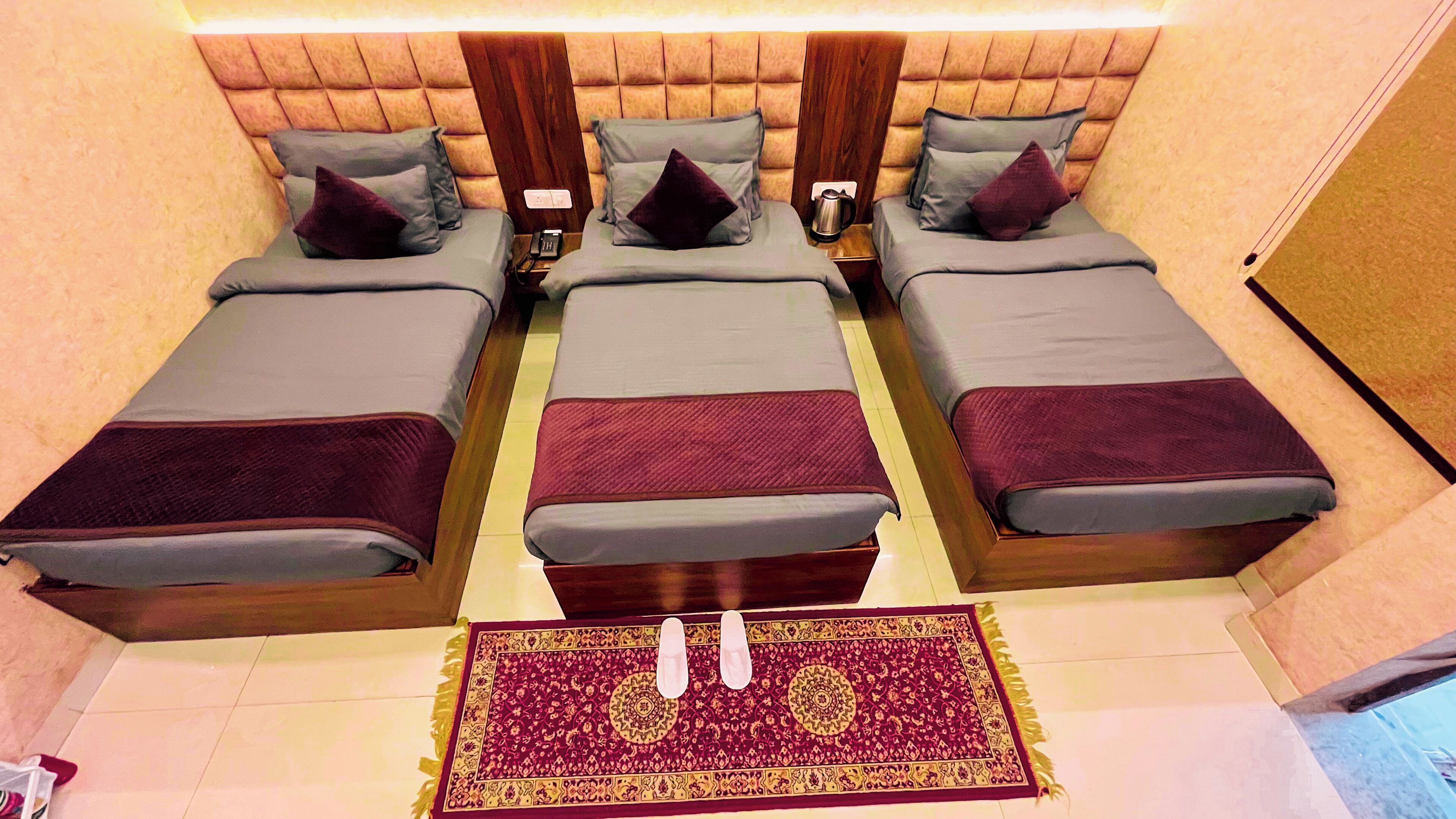 Habitación de hotel con tres camas, cabecera de madera, alfombra estampada y mesita de noche con teléfono y cafetera.