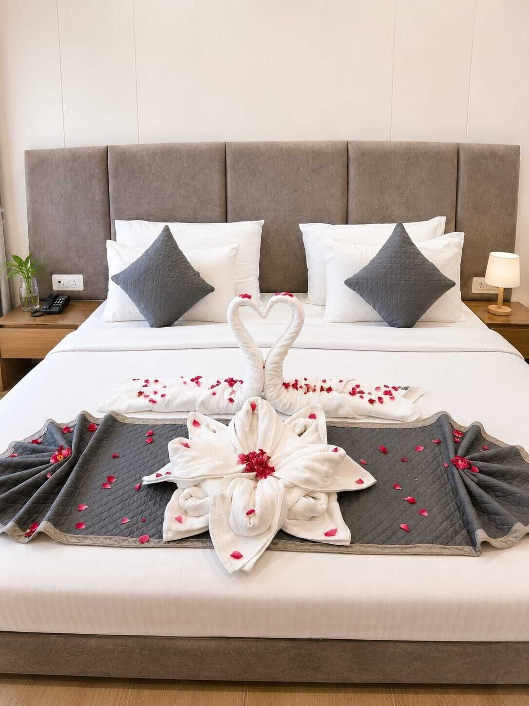 A neatly made bed with a heart-shaped towel arrangement and rose petals, a bedside lamp, and a phone on a bedside table.