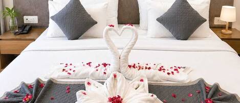 A neatly made bed with a heart-shaped towel arrangement and rose petals, a bedside lamp, and a phone on a bedside table.
