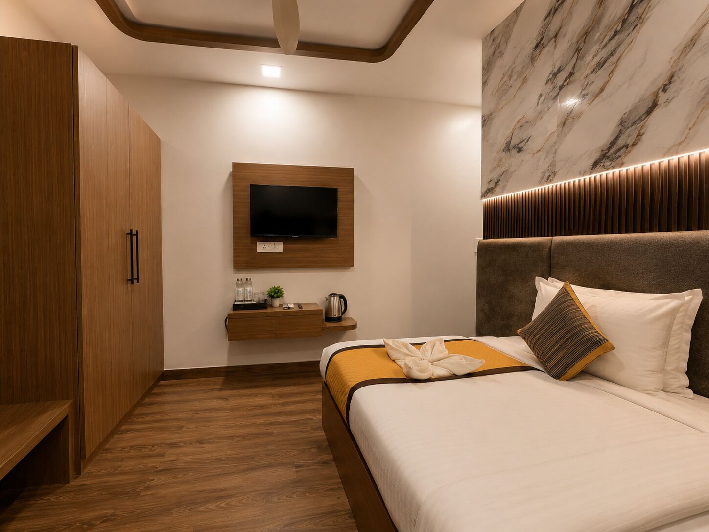 A modern hotel room with a wooden wardrobe, a flat-screen TV mounted on the wall, a bedside table with a kettle, and a bed with a headboard and pillows.