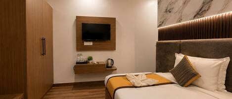 A modern hotel room with a wooden wardrobe, a flat-screen TV mounted on the wall, a bedside table with a kettle, and a bed with a headboard and pillows.