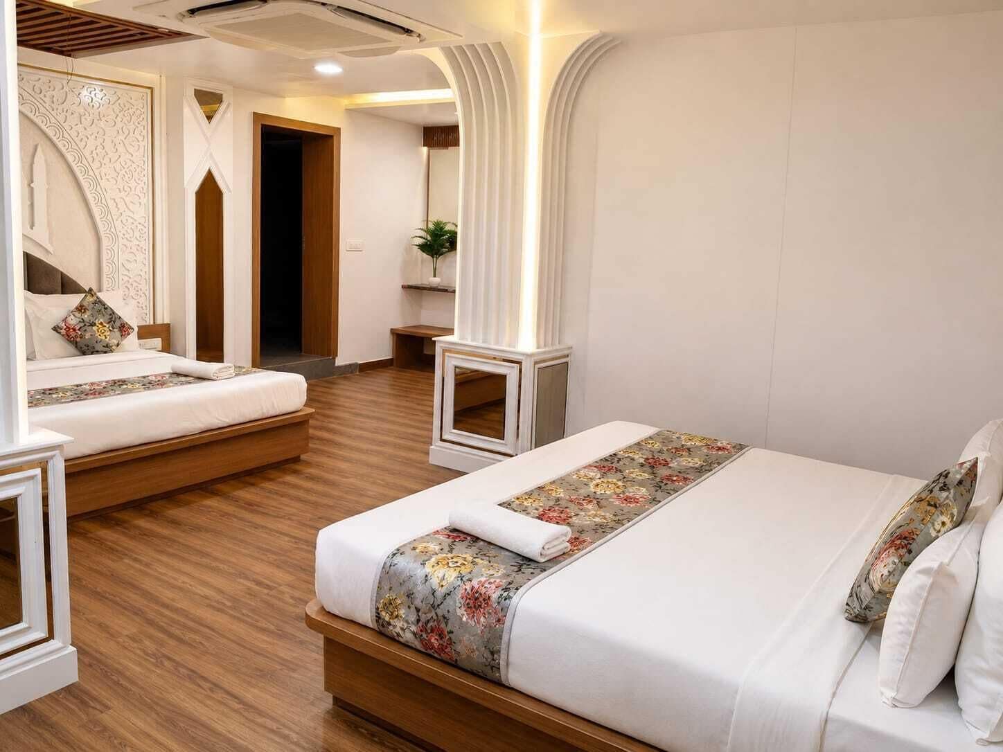 A hotel room with a wooden floor, two beds with floral bedspreads, a mirror, and a small plant.