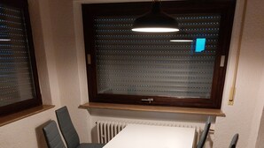 Et spiseområde med et hvidt bord, fire stole, et vindue med skodder og en hængende lampe.