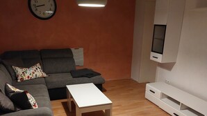 En stue med en grå sofa, et hvidt sofabord, et ur på væggen og et fjernsyn monteret på væggen.