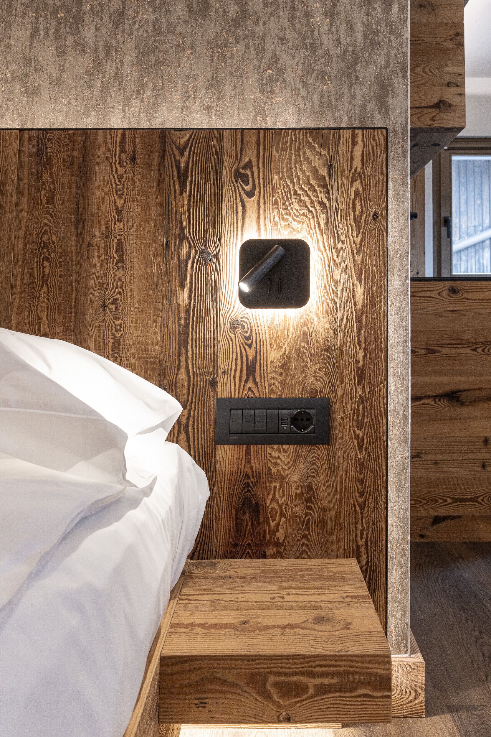 A wooden bed with a nightstand, a wall-mounted light, and a power outlet.