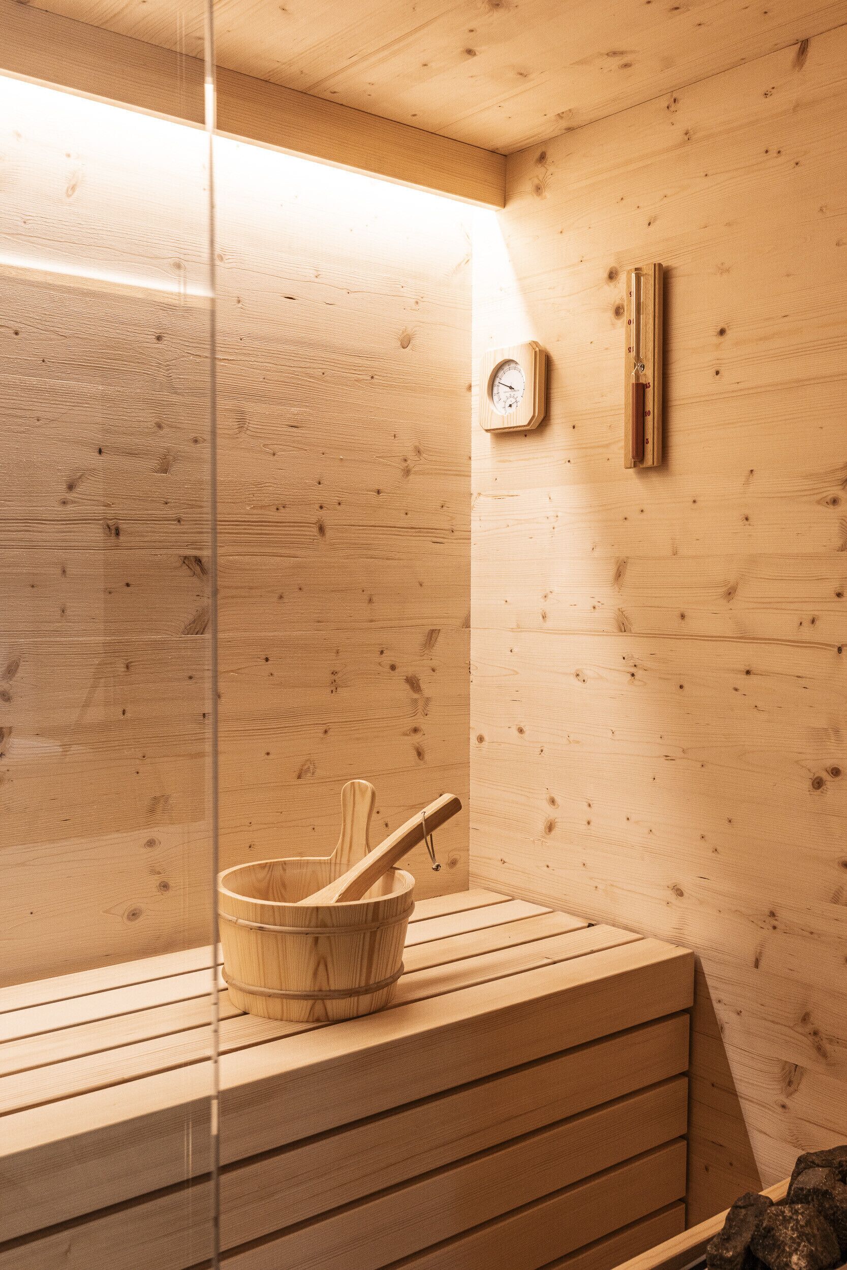 Una sauna de madera con un banco de madera, un cubo de madera y un mazo, y un panel de control de la sauna fijado a la pared.