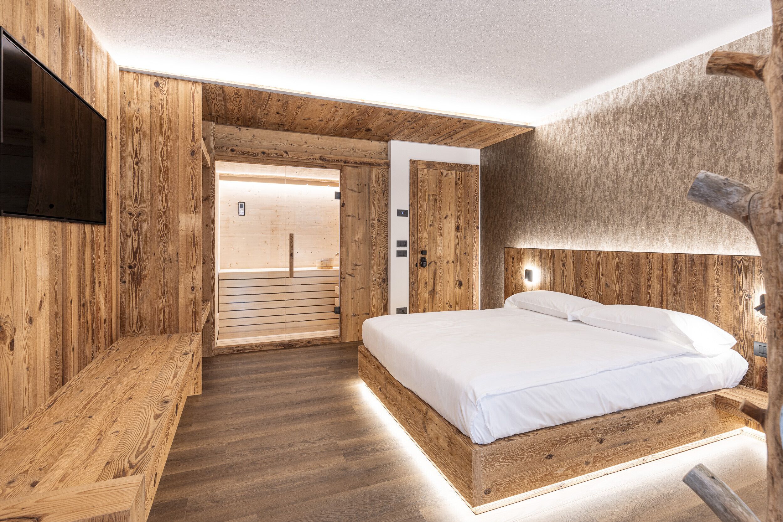 A wooden-themed bedroom with a bed, bench, and a TV mounted on the wall.