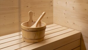 Eine Holzsauna mit einer Holzbank, einem Holzfass mit einem Wasserschöpfe und einem an der Wand befestigten Thermometer.