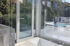 Ein modernes Badezimmer mit Duschkabine aus Glas, einer Badewanne und Blick auf ein Schwimmbecken und die umliegende Natur.