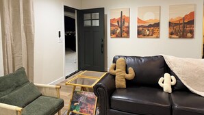 Ein Wohnzimmer mit einem grünen Sessel, einer schwarzen Ledersofa mit Kaktuspflanzenkissen, einem Glastisch und Wandkunst mit Wüstenmotiven.