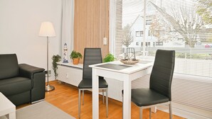 Un salon moderne avec une table blanche, deux chaises noires, un canapé noir, un lampadaire et une vue dégagée grâce à de grandes fenêtres.