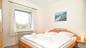 Et soveværelse med en træseng, hvidt sengetøj, et vindue med gardiner, en radiator og et maleri af en strand.