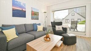En moderne stue med en grå sofa, et træbord og en grå lænestol. Der er et stort vindue med udsigt til en bygning og et landskabsmaleri på væggen.