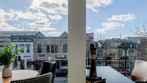 Une vue depuis une fenêtre donnant sur une rue où se trouvent divers commerces, notamment une boulangerie et un restaurant proposant des hamburgers.