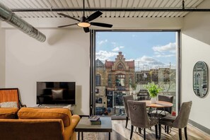 Uma sala de estar moderna com uma janela ampla, uma TV de tela plana, um ventilador de teto e um espaço de jantar com cadeiras e uma mesa.