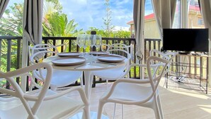 Un área de comedor al aire libre con techo, una mesa dispuesta para dos personas, sillas blancas y un televisor sobre un soporte a la derecha.