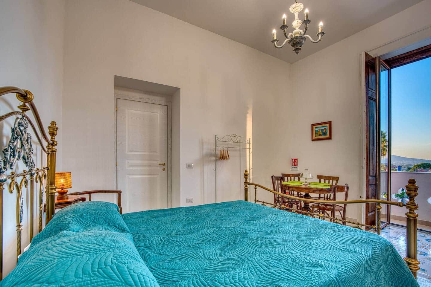 Une chambre à coucher avec un lit, un lustre, une table à manger avec des chaises, et une vue sur l’extérieur par une porte ouverte.
