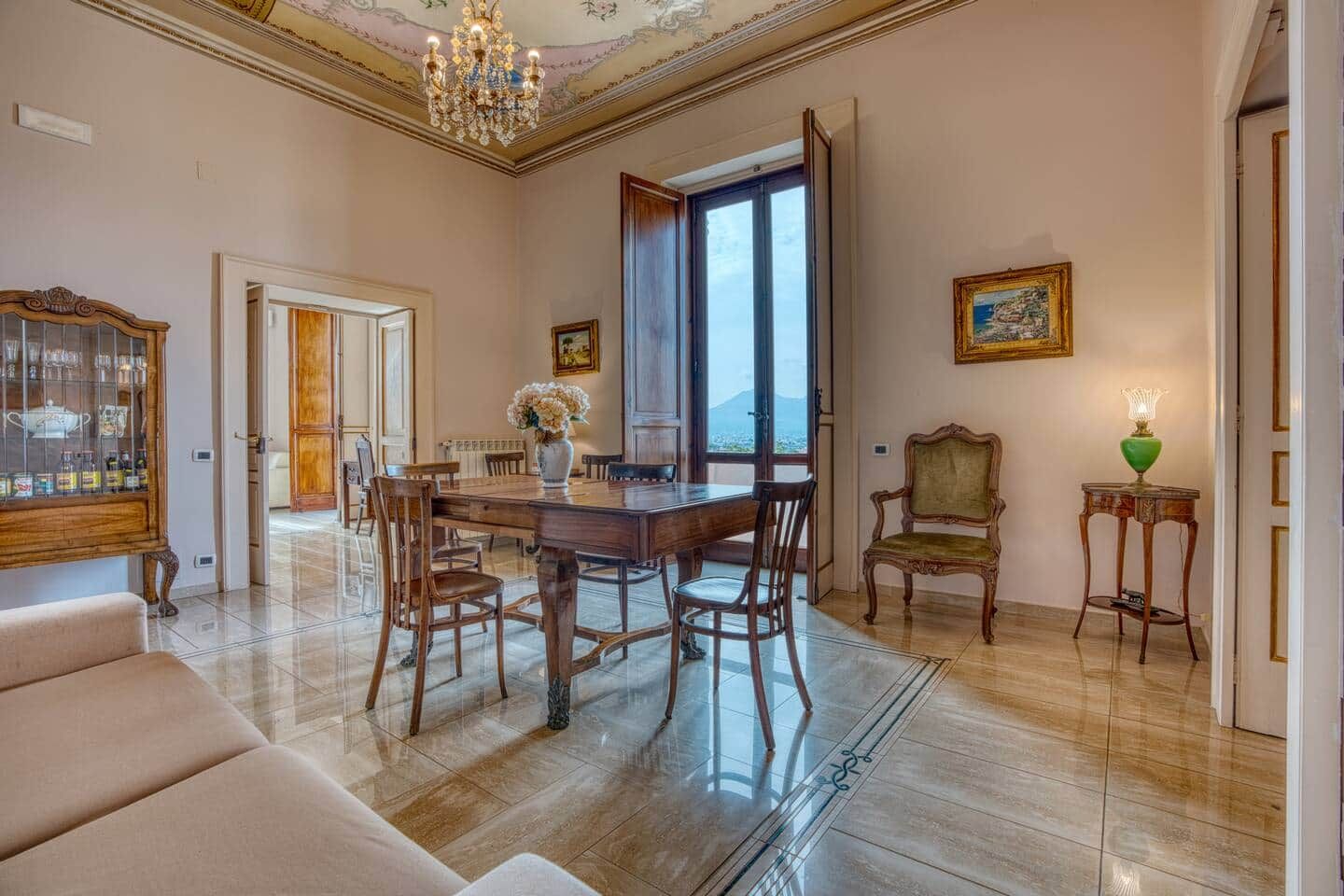 Amplia habitación con una mesa de comedor de madera, sillas, un sofá, una araña y vistas a través de un ventanal.