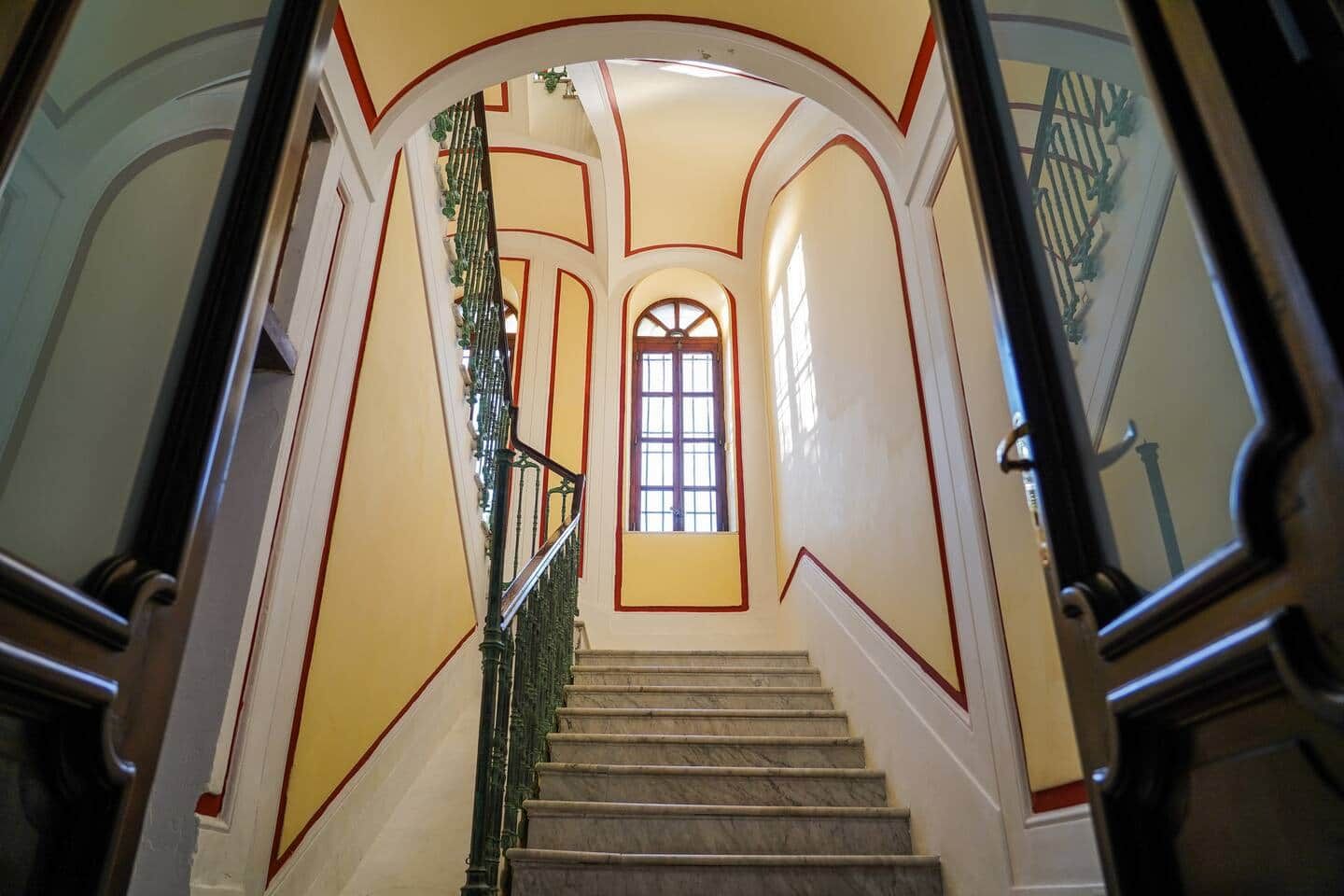 Una imponente escalera con detalles de mármol, barandillas ornamentales y un ventanal grande en la parte superior.