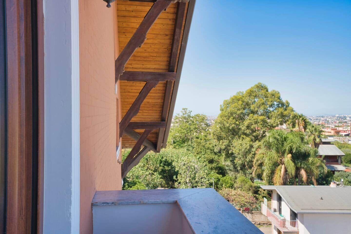Un balcon doté d’une structure en poutres de bois, offrant une vue sur une zone densément végétalisée, avec des arbres et des bâtiments visibles au loin.