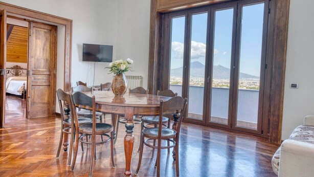 A dining area with a wooden table and chairs, a vase with flowers, and a large window with a view of a mountain and city. - Historic Villa Apartment with Panoramic Views of Mount Vesuvius (Angri)
