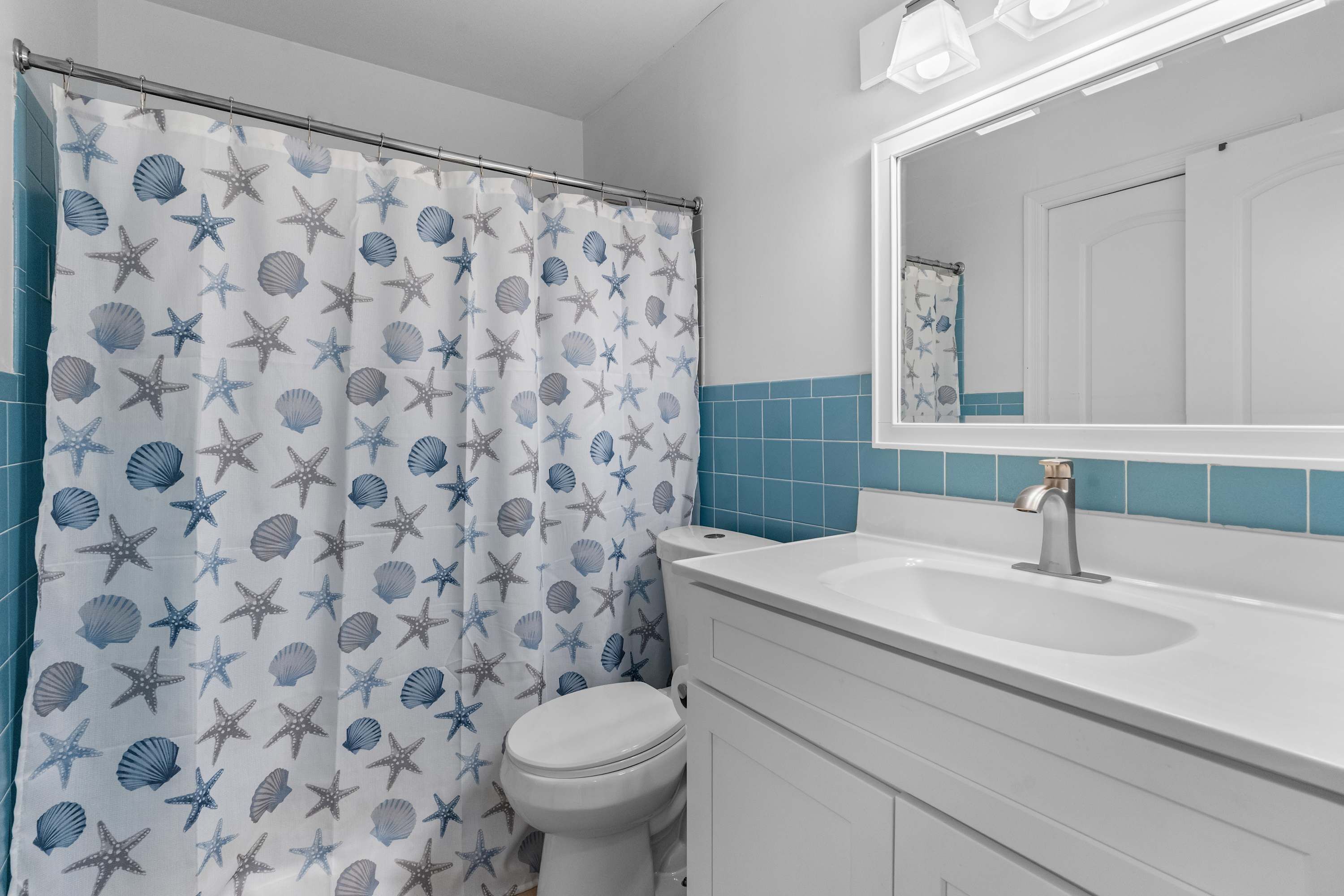 A bathroom with a shower curtain featuring a starfish and seashell pattern, a white toilet, a white sink with a mirror above it, and blue-tiled walls.