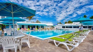 Zona de piscina con sillones de descanso y sombrillas, una piscina y un edificio de fachada blanca con detalles azules.