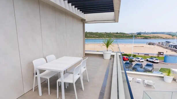 A balcony with a white table and chairs, a potted plant, and a view of a beach and parked cars. - First A - La Torre de Suances (Suances)
