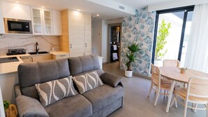 A modern living room with a grey sofa, a dining table with wicker chairs, a kitchen with white cabinets, a microwave, and a potted plant. - First A - La Torre de Suances (Suances)