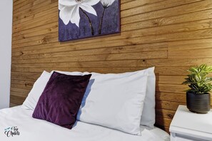 A bed with white and purple pillows, a wooden headboard, a wall-mounted painting of flowers, and a potted plant on a bedside table.