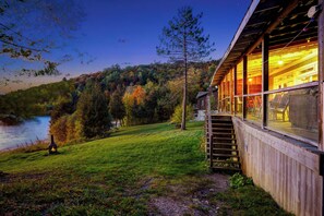 Una cabaña de madera con paredes de vidrio, una terraza de madera y vista a un lago rodeado de árboles.