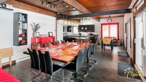 A spacious dining area with a long wooden table set for a meal, surrounded by modern chairs. A kitchen area with a red accent wall and a clock is visible in the background. - LAKE Simon, SPA, private BEACH, 4 bedroom, 2 private office, 10 beds, 16 people. (Lac-Simon)