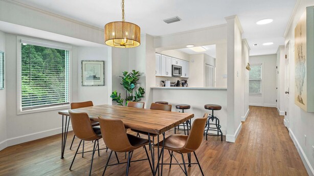 A dining area with a wooden table and chairs, a hanging light fixture, and a view of the kitchen through an open door. - Fly Creek Collection - Bluff View (Fairhope)