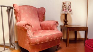 Een hoekzaal met een rode fauteuil met een patroon, een houten bijzettafeltje en een lamp met een bloemenlampenkap.