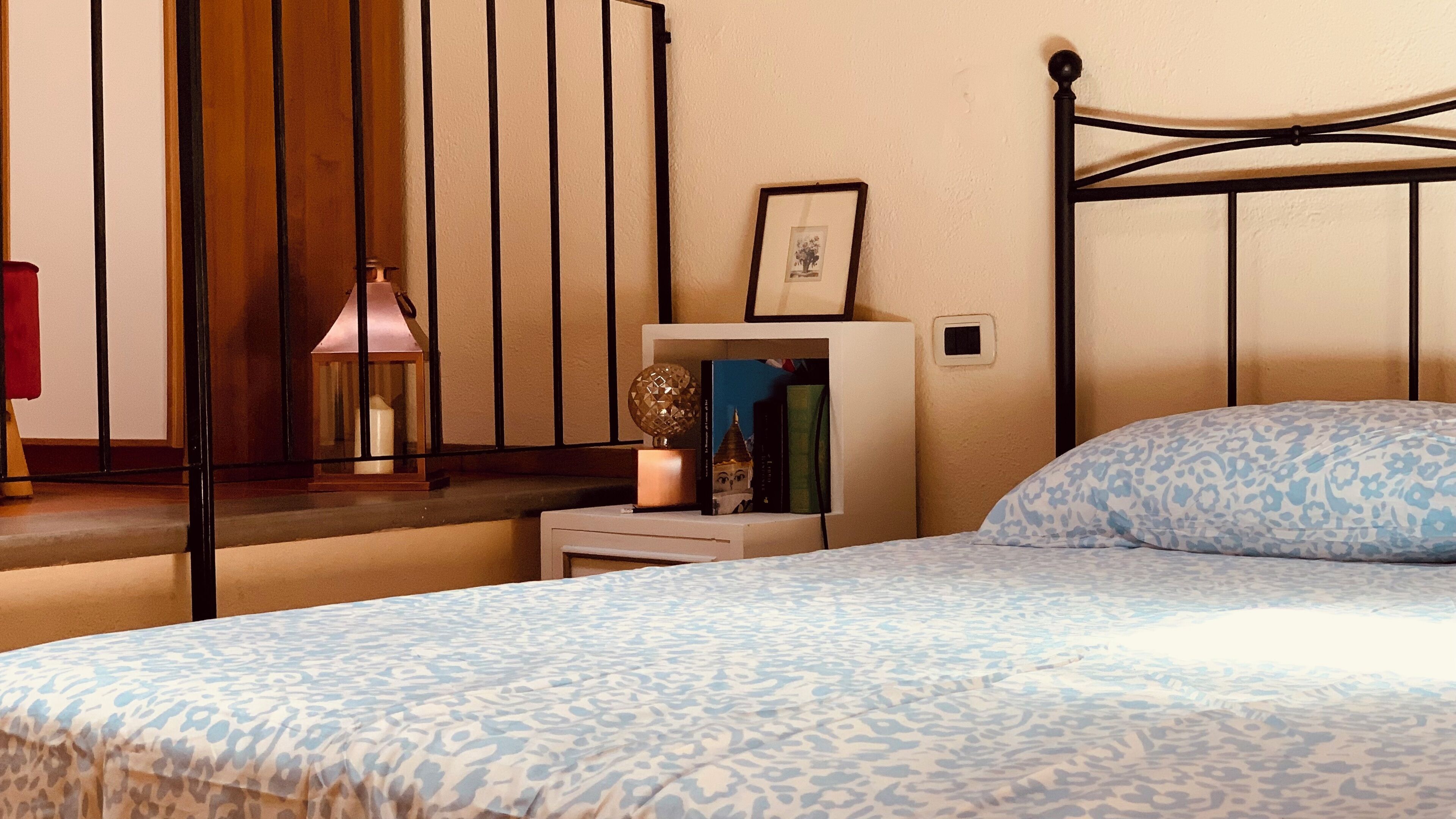 Een slaapkamer met een bed, nachtkastje met lamp, een lijst met afbeelding en een plank met boeken.