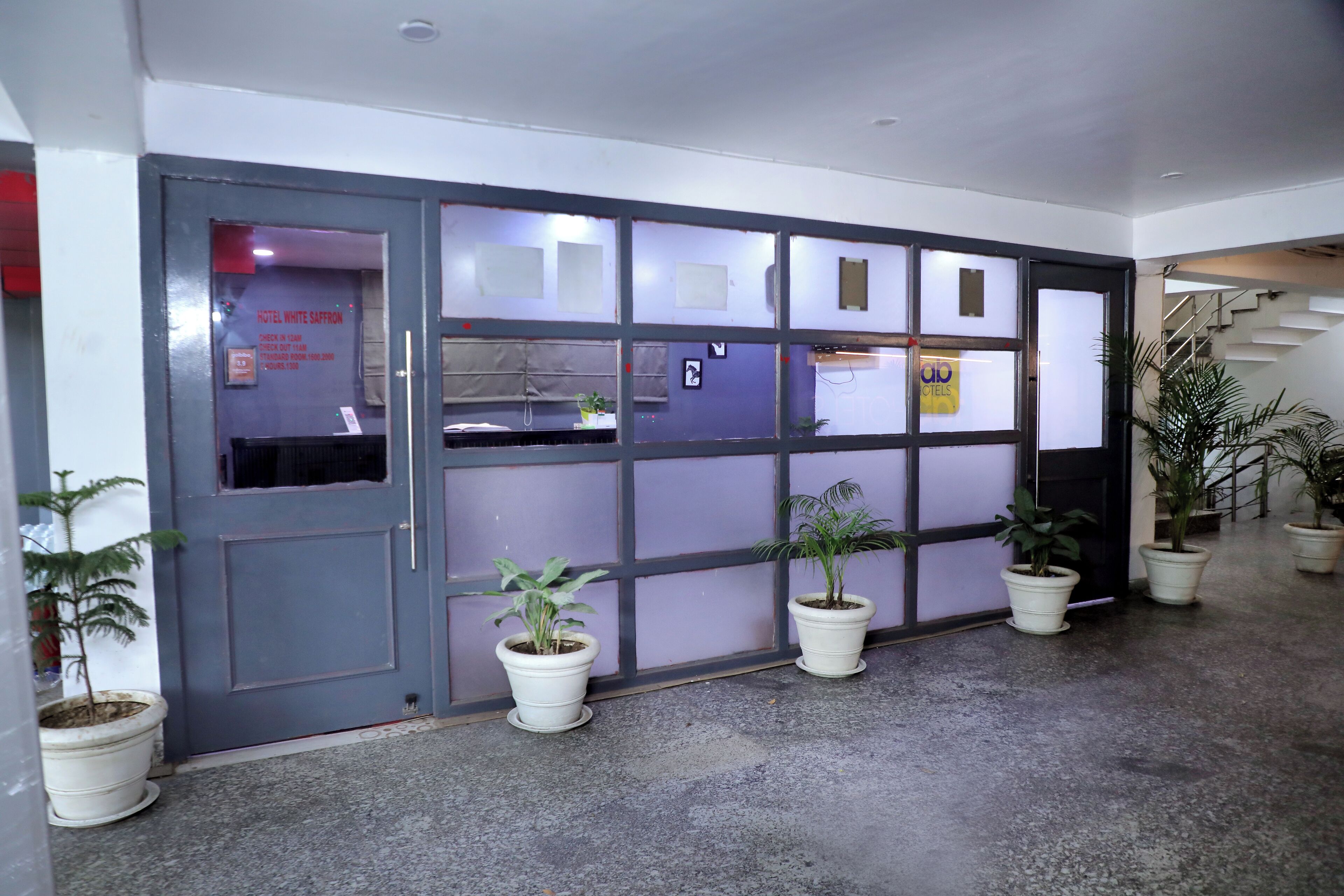 Entrada de hotel com porta cinzenta, várias janelas e plantas em vasos.