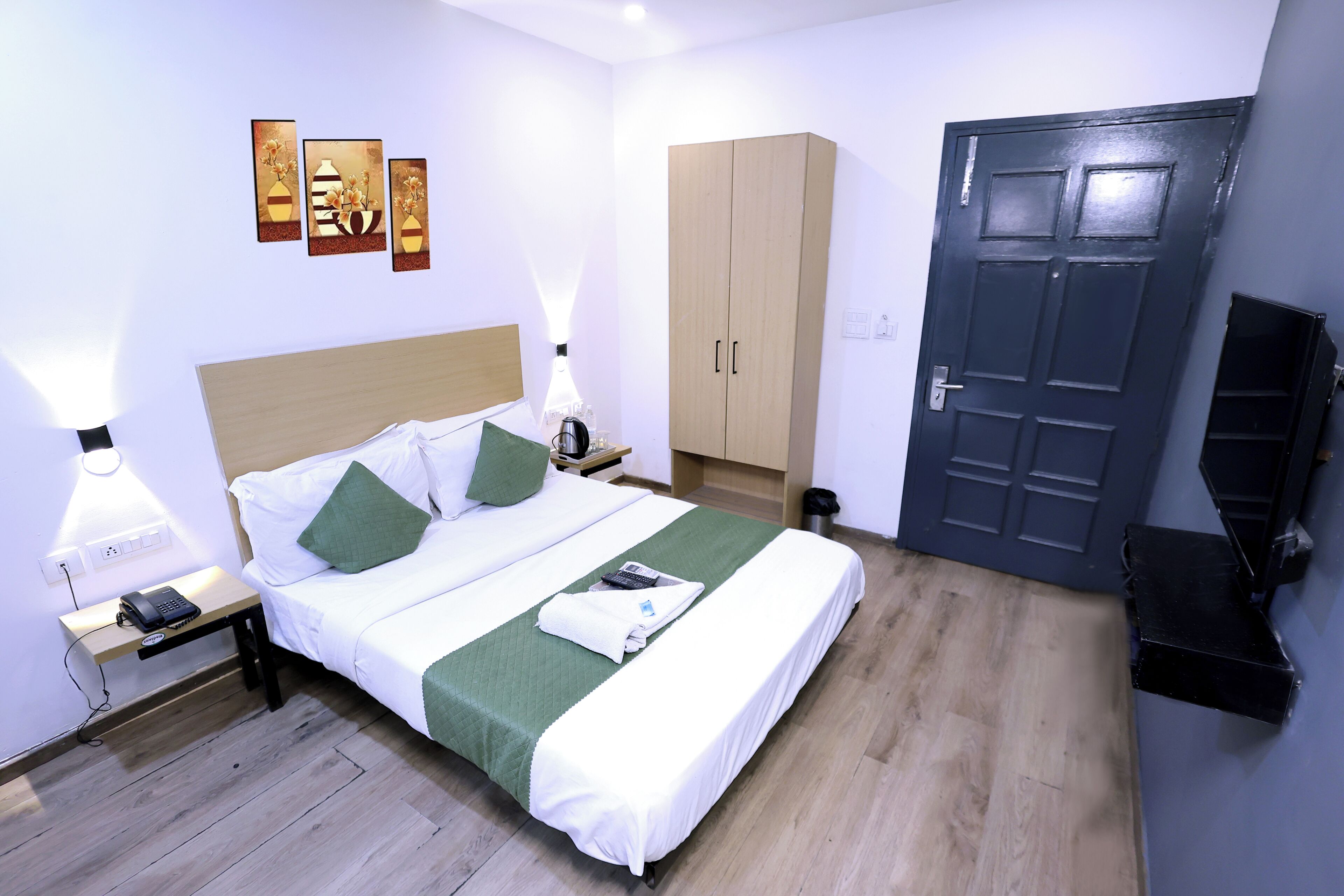 Een netjes ingerichte hotelkamer met een bed, nachtkastje, lamp, wanddecoratie, kledingkast en een deur.