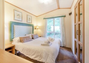 A neatly made bed with pillows and rolled towels, a bedside table, a wall-mounted lamp, and a window with curtains. - 2 bedroom accommodation in Bala, (Bala,)